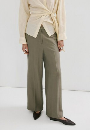 Persona con blusa beige con frente cruzado, pantalones anchos color oliva y zapatos planos negros con punta, de pie contra un fondo blanco liso.