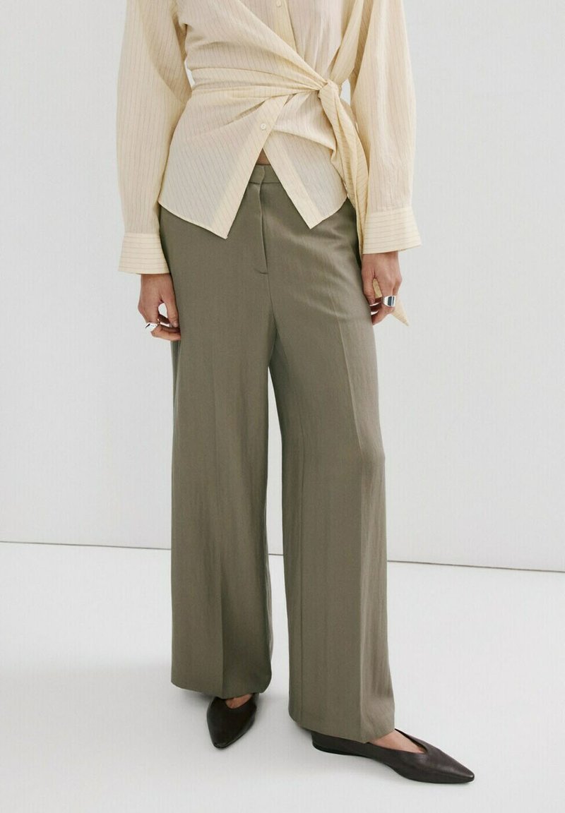 Personne portant un chemisier beige à devant noué, un pantalon large olive et des ballerines noires à bout pointu, debout devant un fond blanc uni.