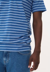 Mėlynos ir šviesiai mėlynos juostelės marškinėliai, trumpomis rankovėmis, laisvas kirpimas, pagaminti iš medvilnės, derinami su mėlynais džinsais. Etiketė matoma apdailoje.