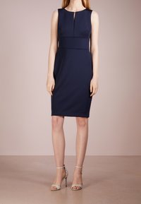 Marineblauwe mouwloze jurk met een keyhole-decolleté, getailleerde pasvorm en gladde stof. Gepaard met lichtgrijze hakken sandalen.