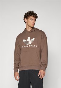 Rudas džemperis su kengūros kišene, ant kurio yra baltas Adidas logotipas ir užrašas "KREPŠINIS". Laisvas siluetas, su įribuotomis rankovėmis ir apačia.