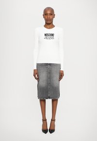 Bela dolga majica z dolgimi rokavi in črnim napisom "MOSCHINO JEANS", v kombinaciji s sivo jeans krilom v obliki svinčnika in črnimi čevlji s konico.