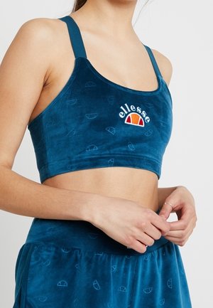 Kobieta w niebieskim sportowym biustonoszu ellesse i dopasowanych niebieskich aksamitnych spodniach, ręce splecione na talii, jasne tło.