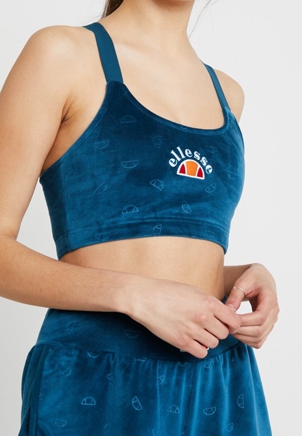 Femme portant un soutien-gorge de sport ellesse bleu et un pantalon en velours bleu assorti, mains jointes à la taille, fond clair.