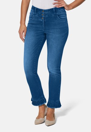 Jean en denim bleu à la coupe cintrée, avec une taille haute, une fermeture à trois boutons et des accents de ourlet plissé. Le mannequin porte des chaussures beiges.
