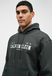 Hoodie grigio scuro con tasca a marsupio frontale, testo bianco a contrasto "CALVIN KLEIN" con data di fondazione, e lacci con estremità metalliche.