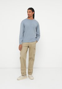 Šviesiai mėlyna ilgomis rankovėmis marškinėliai, šviesiai rudos spalvos kargo kelnės su keliais kišenėmis ir beveik baltos spalvos sportiniai bateliai. Paprastas dizainas, kasdieniškas siluetas, minimalūs detalės.