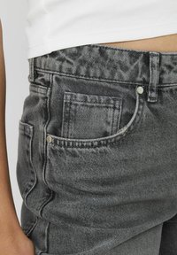 Jean en denim gris avec une texture légèrement décolorée, doté d'une poche avant et d'une fermeture à bouton en argent à la taille.