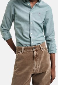 Homme portant une chemise bleu clair à boutons aux manches retroussées, rentrée dans un pantalon marron taille haute, les mains partiellement dans les poches, sur fond blanc.