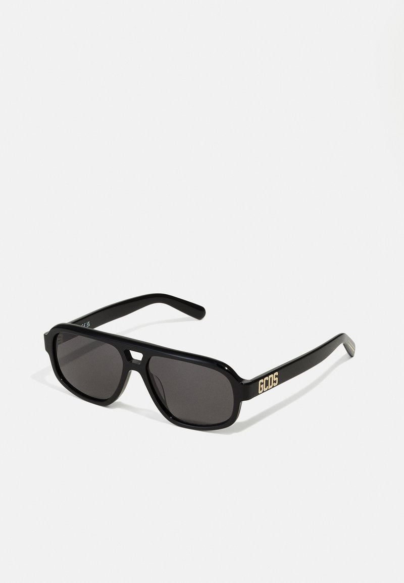 Black sunglasses with dark lenses, thick frames, and a rectangular shape. Features a gold "GCDS" logo on the side. Smooth texture.