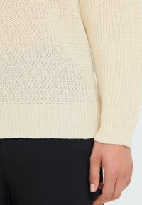 Strickpullover in Creme mit einem strukturierten Muster, kontrastierendem geripptem Saum und engen Bündchen. Getragen mit schwarzen Stoffhosen.