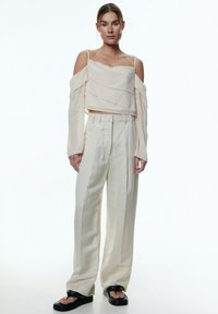Hellbeige cropped Top mit drapierten Lagen, kombiniert mit lockeren, cremefarbenen Hosen. Das Model trägt schwarze Plateausandalen. Insgesamt glatte Textur.