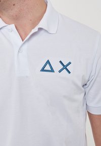 Polo bianco con tessuto testurizzato, caratterizzato da forme geometriche ricamate blu: un triangolo e una 'X' nella zona sinistra del petto.