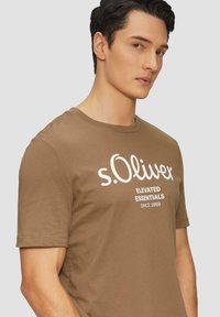 Hnědé bavlněné tričko s krátkým rukávem, na kterém je na hrudi bílý nápis "s.Oliver Elevated Essentials Since 1969".
