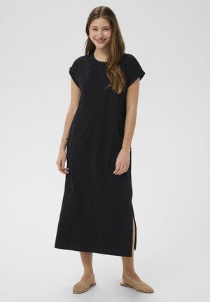 Mladá žena sa usmieva, oblečená v čierne midi šaty s krátkym rukávom a bočným rozparkom a béžové pletené baleríny, stojaca na jednoduchom pozadí.