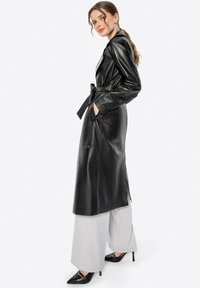 Vrouw in een zwarte leren trenchcoat met riem, witte wijde broek en zwarte puntige hakken, staand tegen een effen achtergrond.