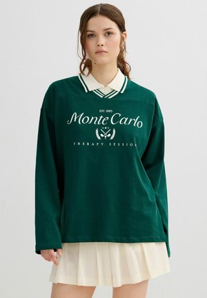 Mujer joven con cabello castaño, vestida con una camisa de manga larga verde oscuro con la inscripción "Sesión de Terapia Monte Carlo" sobre una blusa blanca con cuello y falda plisada.
