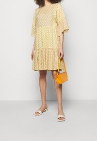Żółta sukienka w wzory z falbaniastym dołem, rozszerzanymi rękawami, z lekkiego materiału. Trzyma małą pomarańczową torbę, a na stopach ma białe klapki.