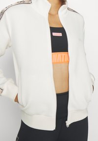 Chaqueta blanca con cremallera, cuello alto, puños elásticos y sujetador deportivo negro de P.E Nation. Presenta un logo en contraste y ribete estampado a lo largo de los hombros.