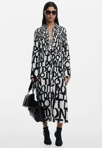 Schwarz-weißes Hemdkleid mit großen Schriftzügen, langen Ärmeln und tailliertem Gürtel, kombiniert mit einer schwarzen Handtasche und Stiefeletten.
