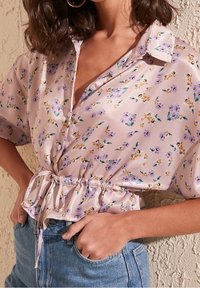 Blouse en satin rose clair avec un imprimé floral, manches courtes, fermeture à boutons à l'avant et taille cintrée nouée avec un ruban.