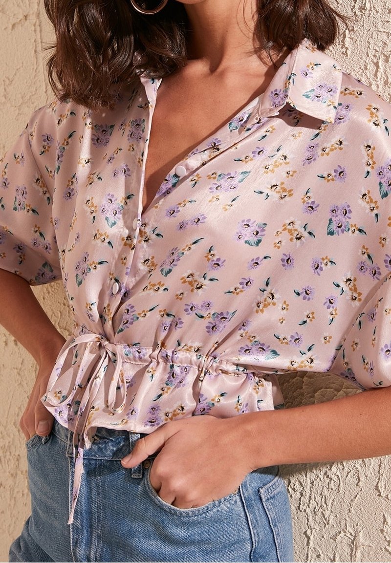 Blouse en satin rose clair avec un imprimé floral, manches courtes, fermeture à boutons à l'avant et taille cintrée nouée avec un ruban.