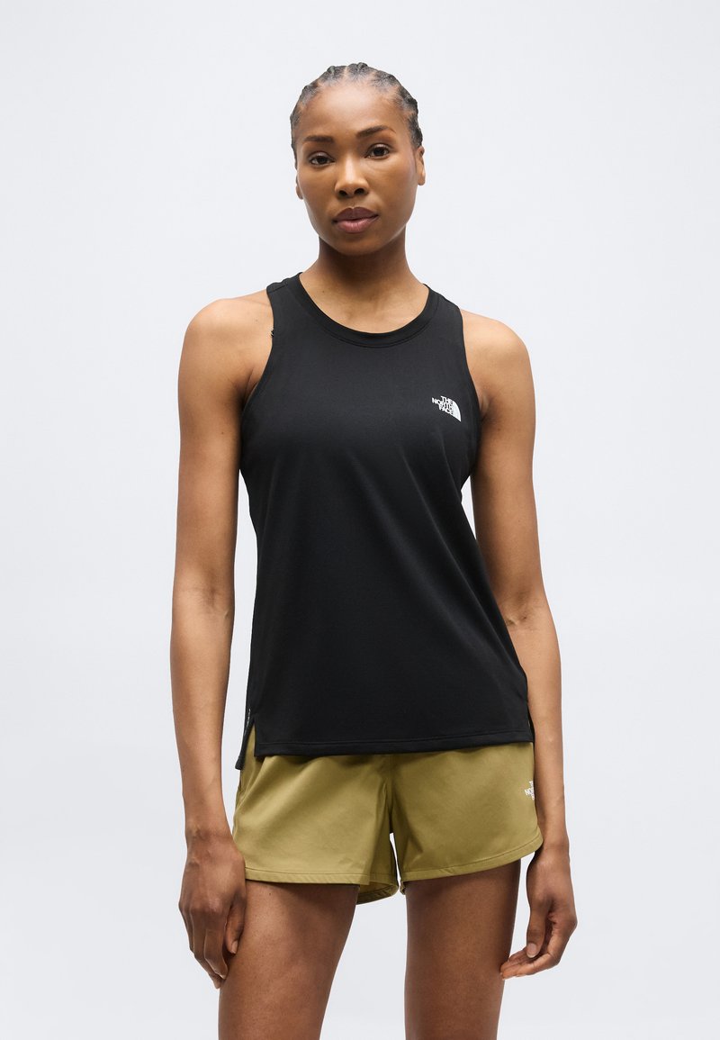 Γυναίκα με πλεγμένα μαλλιά που φοράει μαύρο αμάνικο τοπ και χακί πράσινο σορτς, στέκεται μπροστά σε απλό ανοιχτόφωτο φόντο.