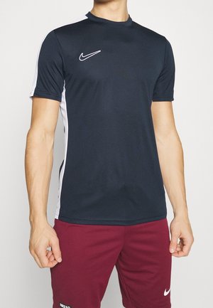 Mężczyzna ubrany w granatową sportową koszulkę Nike z krótkim rękawem i białymi panelami bocznymi oraz bordowe spodenki z białym logo Nike.
