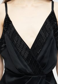 Μαύρο αμάνικο τοπ με βαθύ V-λαιμό, διακοσμημένο με διαγώνιες μαύρες χάντρες κατά μήκος του λαιμού και των ώμων.