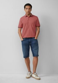 Moška polo majica s kratkimi rokavi v lososovi roza barvi z kontrastnim mornarsko modrim ovratnikom, v kombinaciji z modrimi jeans kratkimi hlačami in bež supergami.