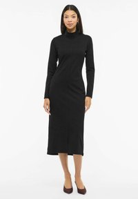 Černé šaty s dlouhým rukávem a rolákem, s přiléhavým střihem a délkou ke kolenům. Hladká látka s detailním středovým švem.