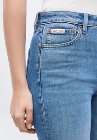 Κοντινό πλάνο ενός ατόμου που φοράει ανοιχτό μπλε τζιν Calvin Klein με χαλκομανίες και λευκό τοπ, το χέρι ακουμπάει στο πλάι.