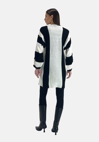 Strik cardigan i sort og hvid. Har brede vandrette og lodrette striber, lange ærmer og en løs pasform. Bæres over sorte leggings og støvler.