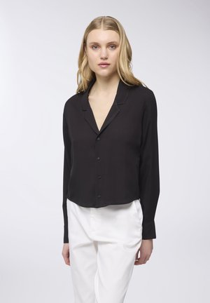 Blusa nera a maniche lunghe con scollatura a V, davanti con bottoni e stoffa morbida. Abbinata a pantaloni bianchi a vita alta.