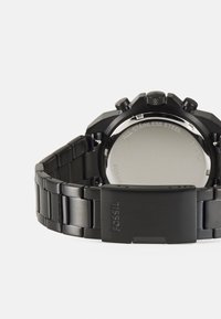 Zwarte roestvrijstalen horloge met een gestructureerde band. Ronde wijzerplaat met chronograaffuncties en een sluitingdetail. Gepolijste achterkant weerkaatst licht.