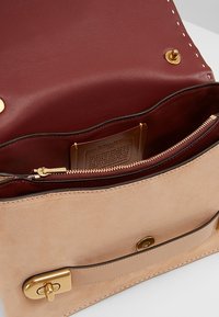 Borsa in suede color cammello con bordi in pelle liscia. L'interno presenta una fodera rosso scuro, hardware dorato e una tasca con zip per una sicura conservazione.