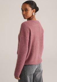 Femme aux cheveux châtains en chignon, portant des boucles d'oreilles en argent, un pull rose duveteux et un jean en denim gris, se retournant par-dessus son épaule.