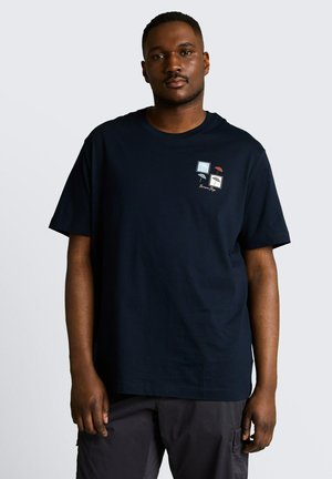 Mężczyzna noszący czarną koszulkę z małymi graficznymi kwadratami i napisem na lewej piersi, w połączeniu z ciemnymi spodniami, stojący na jednolitym tle.