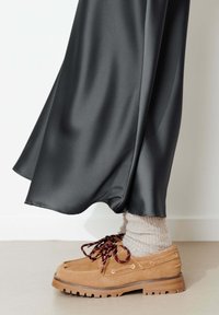Des chaussures en daim marron avec des semelles épaisses et des lacets rouges portées avec des chaussettes beiges sous une jupe noire en satin fluide.