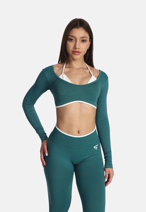 Kvinde iført teal crop top med lange ærmer, matchende højtaljede leggings og hvid bikini top, stående med én hånd på hoften mod en hvid baggrund.