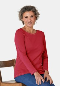 Rode longsleeve shirt met een ronde hals, gemaakt van gladde stof; gecombineerd met blauwe spijkerbroek en een houten stoel op de achtergrond.