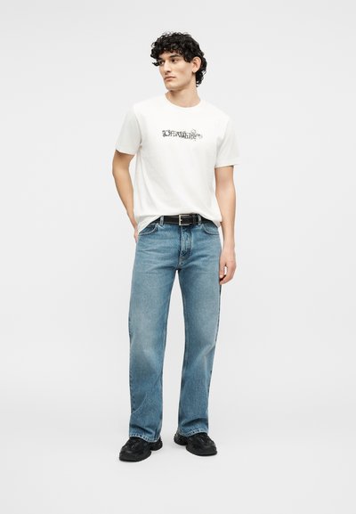 Νεαρός με σγουρά μαλλιά που φοράει λευκό μπλουζάκι με στάμπα, μπλε τζιν, μαύρη ζώνη και μαύρα αθλητικά παπούτσια, στέκεται με το ένα χέρι στην τσέπη.