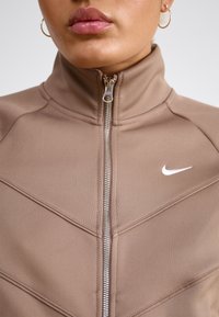 Smeđi kaput na zatvaranje s visokom kragnom, rebrastom teksturom i bijelim Nike logotipom. Ima kutnu šavnu obradu i srebrni zatvarač.