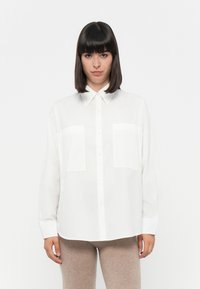 Camicia bianca con bottoni, maniche lunghe, due tasche sul petto e colletto classico. Il tessuto appare liscio con una texture leggera.