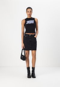 Svart ermeløs crop topp med "TOMMY JEANS" grafikk, sammen med et svart denimskjørt og høye svarte bøyle støvler. Liten svart håndveske.