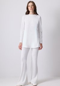 Donna in piedi contro uno sfondo bianco uniforme, vestita con una tunica bianca a maniche lunghe con bottoni laterali e pantaloni bianchi svasati.