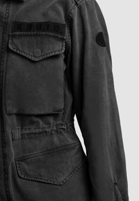 Donkergrijze jas met twee klepzakken aan de voorkant, elastische tailleband, zwarte patch op de borst met letters, en een ronde patch op de mouw.