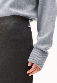 Grijze gebreide trui met ribbels aan de manchetten, gecombineerd met een donkergrijze stoffen rok. Een label is zichtbaar op de zijnaad van de rok.