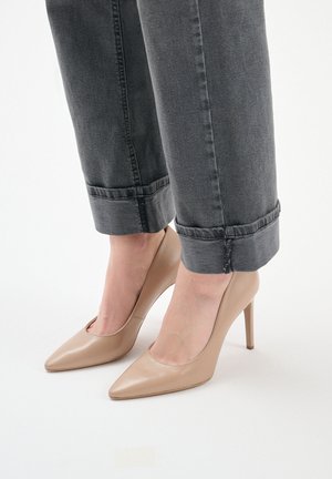 Beżowe lakierowane szpilki z ostrym noskiem, noszone z podwiniętymi ciemnoszarymi dżinsami, podkreślające elegancki design butów.
