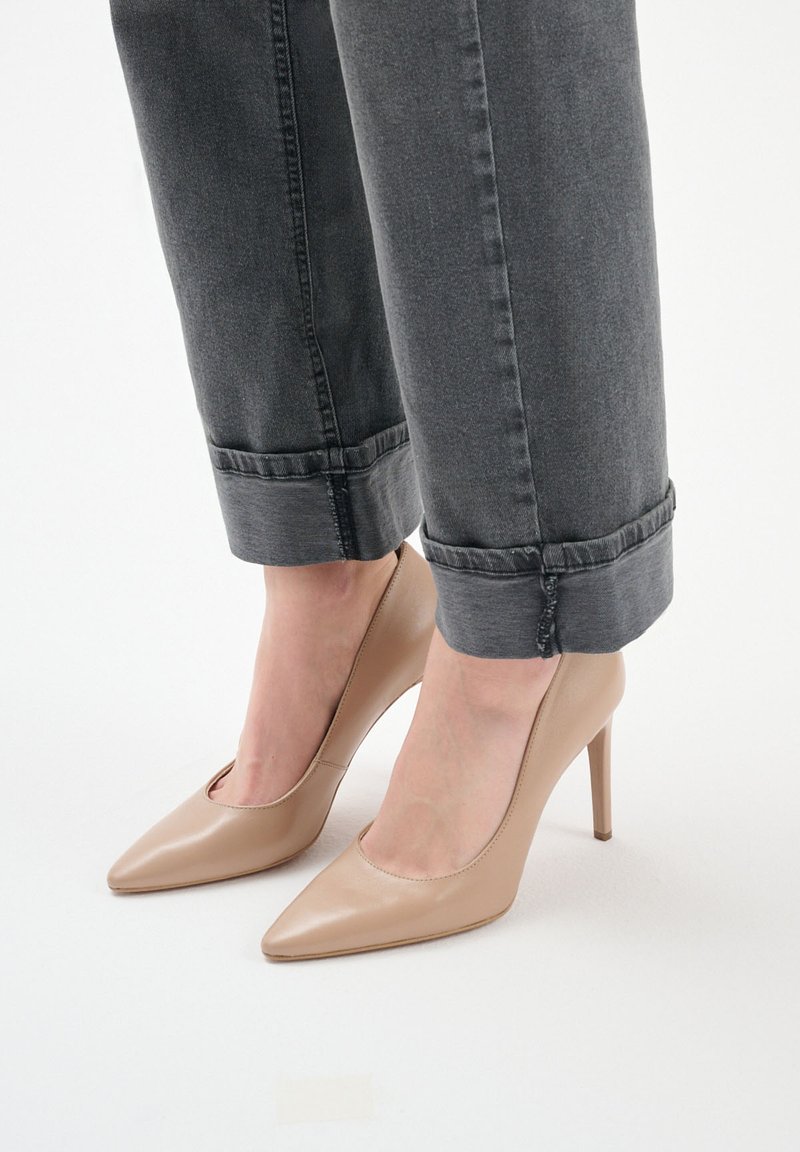 Beżowe lakierowane szpilki z ostrym noskiem, noszone z podwiniętymi ciemnoszarymi dżinsami, podkreślające elegancki design butów.